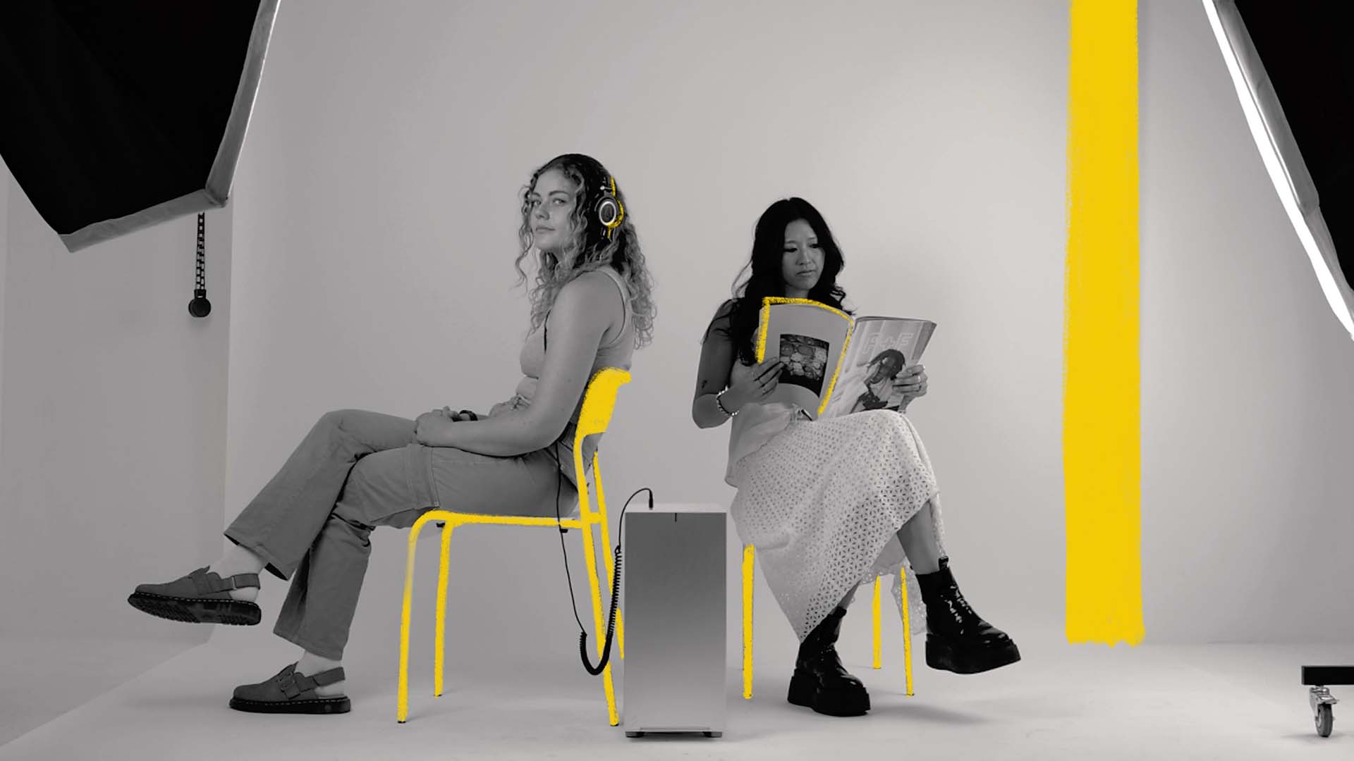 Two creators sitting back-to-back in a minimalist studio, one wearing headphones and the other reading a magazine, with bright yellow highlights on chairs and props beside a Chillblast Spark workstation.
