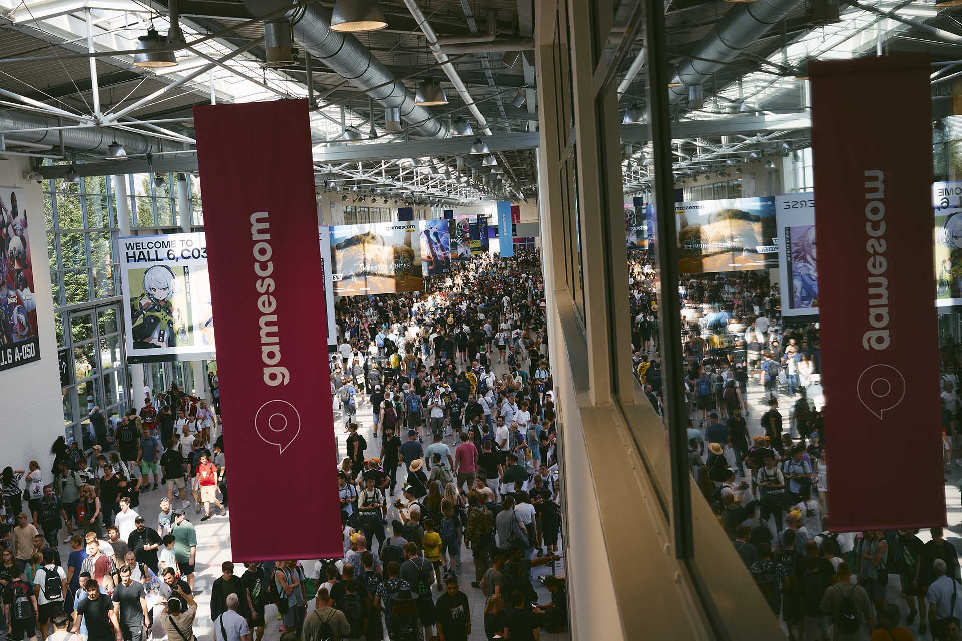 Large crowd of attendees walking through a bright, spacious hall at Gamescom, with banners, posters, and game advertisements hanging overhead.