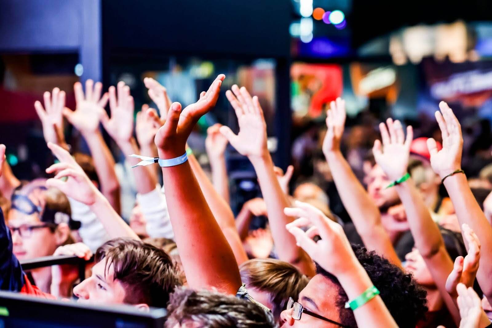 Close-up of a crowd at Gamescom with numerous attendees raising their hands in excitement, wearing event wristbands, and facing a stage or presentation.