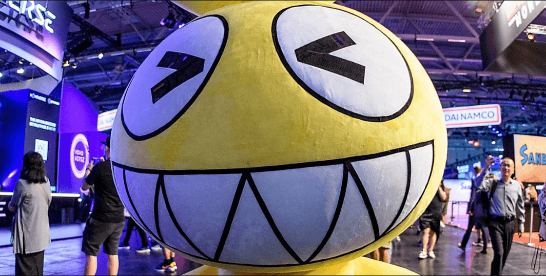 Close-up of a large yellow mascot with exaggerated grinning face and squinted eyes at the Gamescom show floor, with attendees and booths in the background.