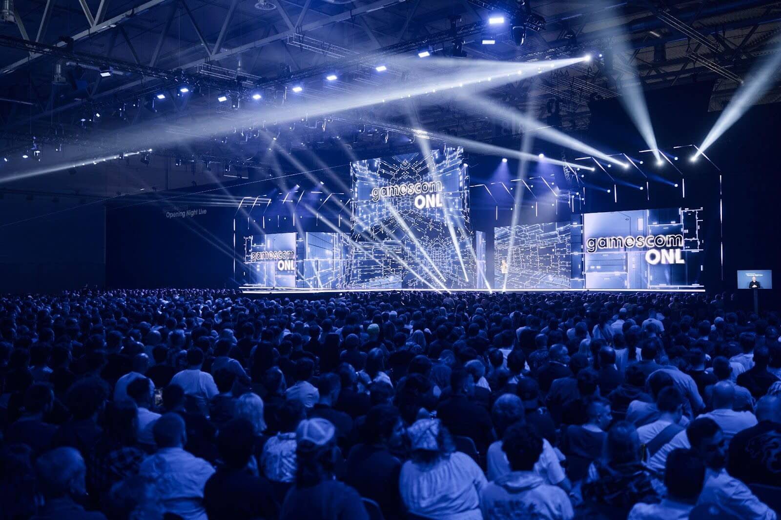 Large audience watching the Gamescom Opening Night Live stage, illuminated with blue lighting, spotlights, and multiple digital screens displaying the event logo.