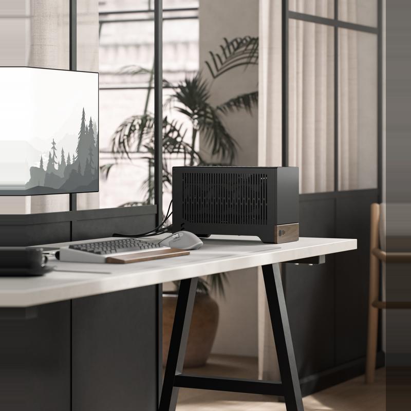 Chillblast Fika computer from the Hej range in Black, with a compact, minimalist design featuring wooden accents, placed on a modern white desk alongside a sleek monitor, keyboard, and mouse, in a bright workspace with large windows and indoor plants.