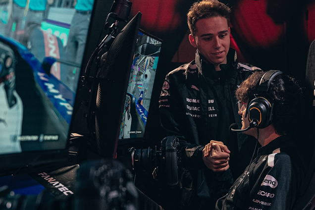 Brown haired man with glasses playing in a racing sim tournament sitting down watching his screen with a headset on, whilst another man with short brown hair stands to his right hand side talking to him 