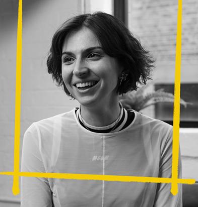 Portrait of a 3D designer with a short haircut and a bright smile, wearing a light sheer top, set in a modern workspace with yellow graphic accents framing the image, representing the creative professionals who use the Chillblast Spark Creator PC range.
