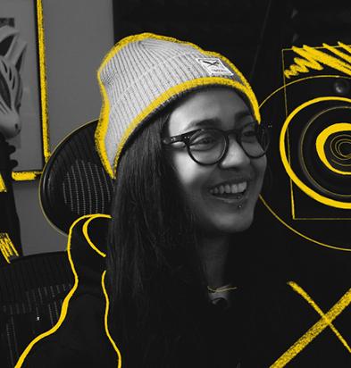 Portrait of a music producer wearing a gray beanie, glasses, and a hoodie, smiling in a creative studio environment with vibrant yellow graphic accents highlighting musical equipment, representing users of the Chillblast Spark Creator PC range.