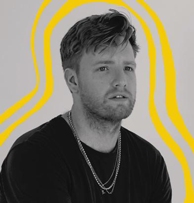 Portrait of a photographer with short hair, wearing layered necklaces and a black shirt, set against a neutral background with vibrant yellow graphic lines, representing creative professionals who use the Chillblast Spark Creator PC range.