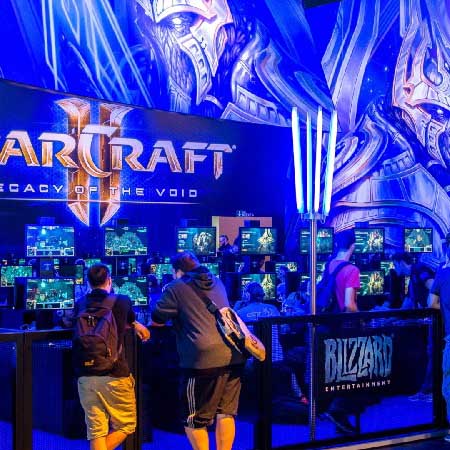StarCraft II: Legacy of the Void booth at a gaming event, with players seated at monitors and large Protoss-themed artwork displayed in the background, hosted by Blizzard Entertainment.